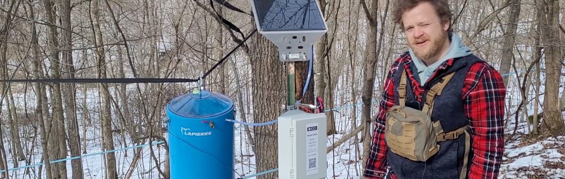 Tony Johnson stands next to the monitoring equipment: sap collection bucket, control panel, solar panel.