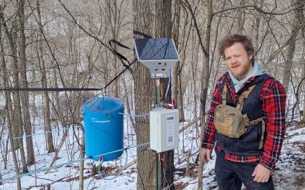 Tony Johnson stands next to the monitoring equipment: sap collection bucket, control panel, solar panel.